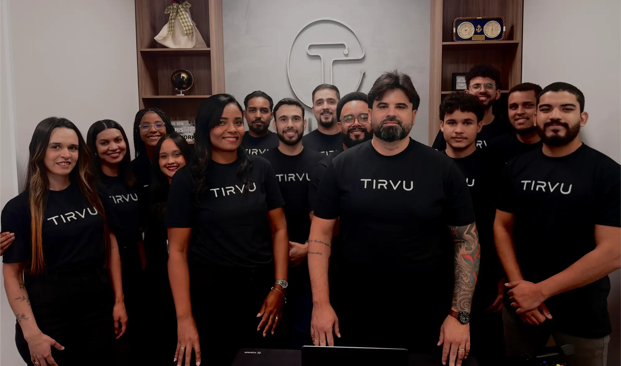 O time da TIRVU reunido no escritório, com camisetas da marca e o logo na parede ao fundo